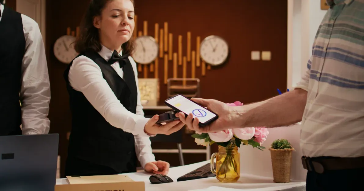 A hotel front desk staff member holds a card machine while a guest pays at the desk.