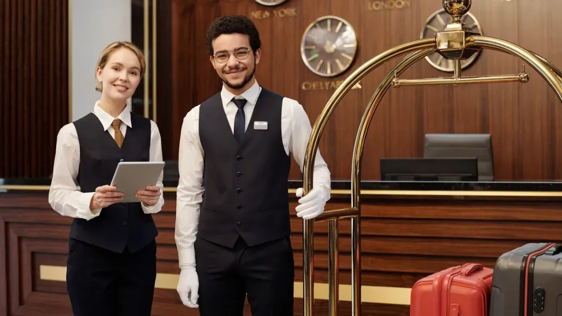 A hotel receptionist and a colleague in uniform stand at the front desk, each holding a tablet.